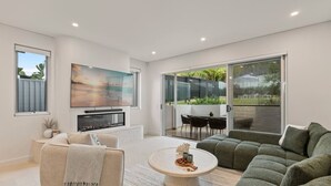 A modern living room with a fireplace, a large TV, a green sofa, and a dining area with outdoor views. - Bayside Luxury Retreat with Pool Near to Cronulla (Port Hacking)