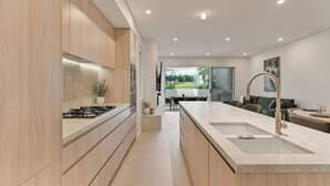 A modern kitchen with a central island, a dining area with a table and chairs, and a view of the outdoors. - Bayside Luxury Retreat with Pool Near to Cronulla (Port Hacking)