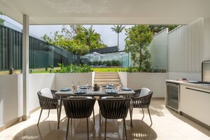 A covered outdoor dining area with a table set for four, surrounded by wicker chairs, and a kitchen area to the right.