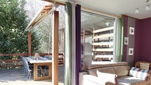 A room with a wooden deck, a sofa, a dining table with chairs, and a view of trees.