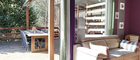 A room with a wooden deck, a sofa, a dining table with chairs, and a view of trees.