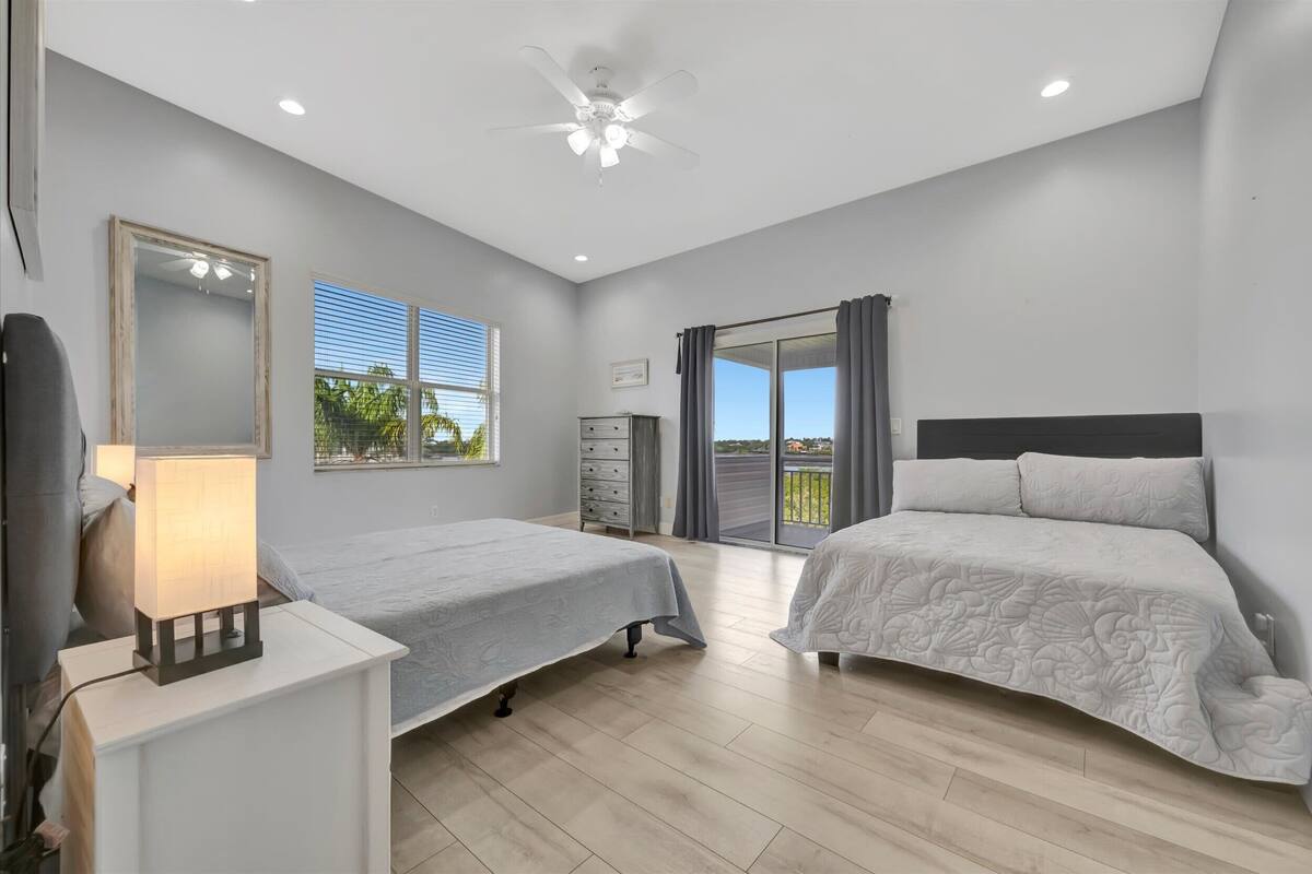 Une chambre avec deux lits, un ventilateur de plafond, une table de chevet avec une lampe, une commode et une grande fenêtre avec des stores.