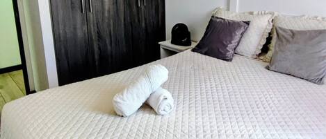 A neatly made bed with a quilted white duvet, a dark wooden wardrobe, and a nightstand with a black bag.