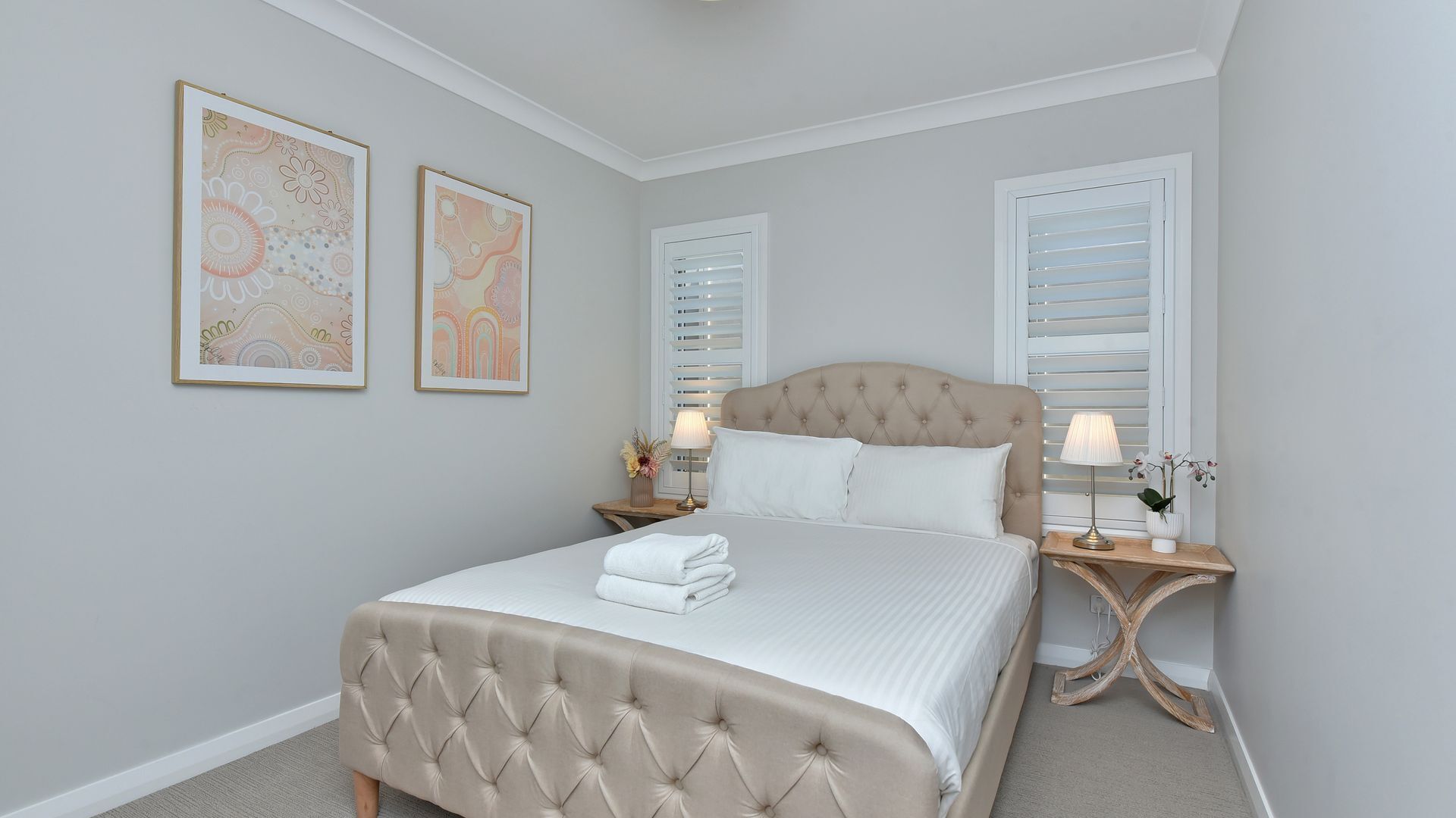Une chambre à coucher avec une tête de lit capitonnée, du linge de lit blanc, deux tables de chevet avec des lampes et deux œuvres d’art abstraites encadrées accrochées au mur.