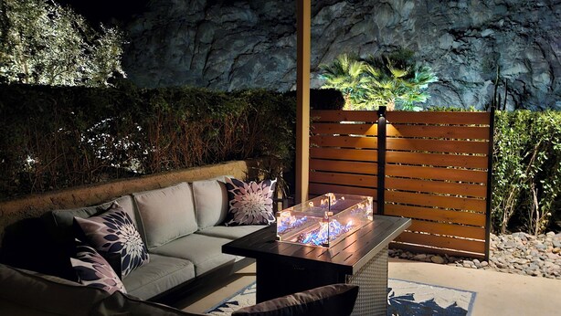Una moderna zona de estar al aire libre con un sofá, cojines, una mesa con una chimenea y un muro de rocas de fondo.