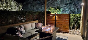 Una moderna zona de estar al aire libre con un sofá, cojines, una mesa con una chimenea y un muro de rocas de fondo.