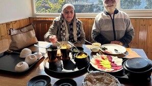 Dos personas sentadas a una mesa con diversos platos, incluyendo sopa y carne, en una habitación con un ventanal que ofrece vistas a un paisaje montañoso.