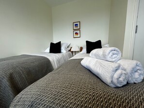 Quarto de hotel com duas camas de solteiro, uma mesa de cabeceira com abajur e uma porta.