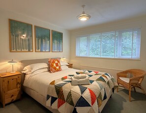 A bedroom with a bed, a nightstand, a chair, a window with blinds, and three paintings on the wall. - 16 Manesty View -  a bungalow that sleeps 4 guests  in 2 bedrooms (Central Keswick)