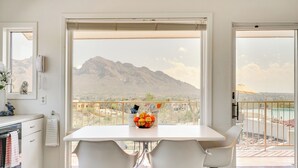 Un coin repas comprenant une table, des chaises, un panier de fruits et une vue sur les montagnes, visible à travers de grandes fenêtres.