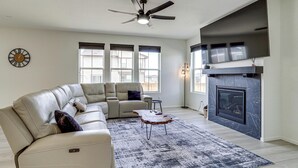 A living room with a sectional sofa, a fireplace, a ceiling fan, and large windows.