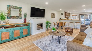Ein Wohnzimmer mit Fernseher, Holzboden, einem Essbereich mit Stühlen, einer Küchenzeile und einem großen Spiegel an der Wand.