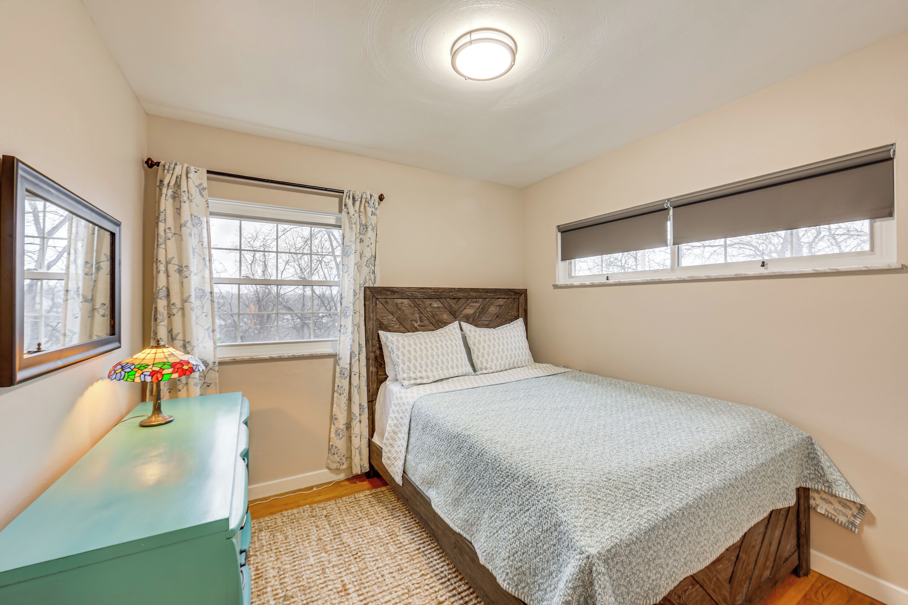 A bedroom with a bed, a dresser, a lamp, a window with curtains, and a ceiling light.