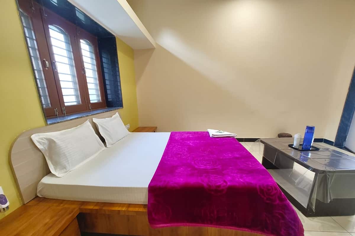Quarto com uma cama, uma mesa de cabeceira de madeira, uma pequena mesa com um frasco azul e uma janela com persianas.