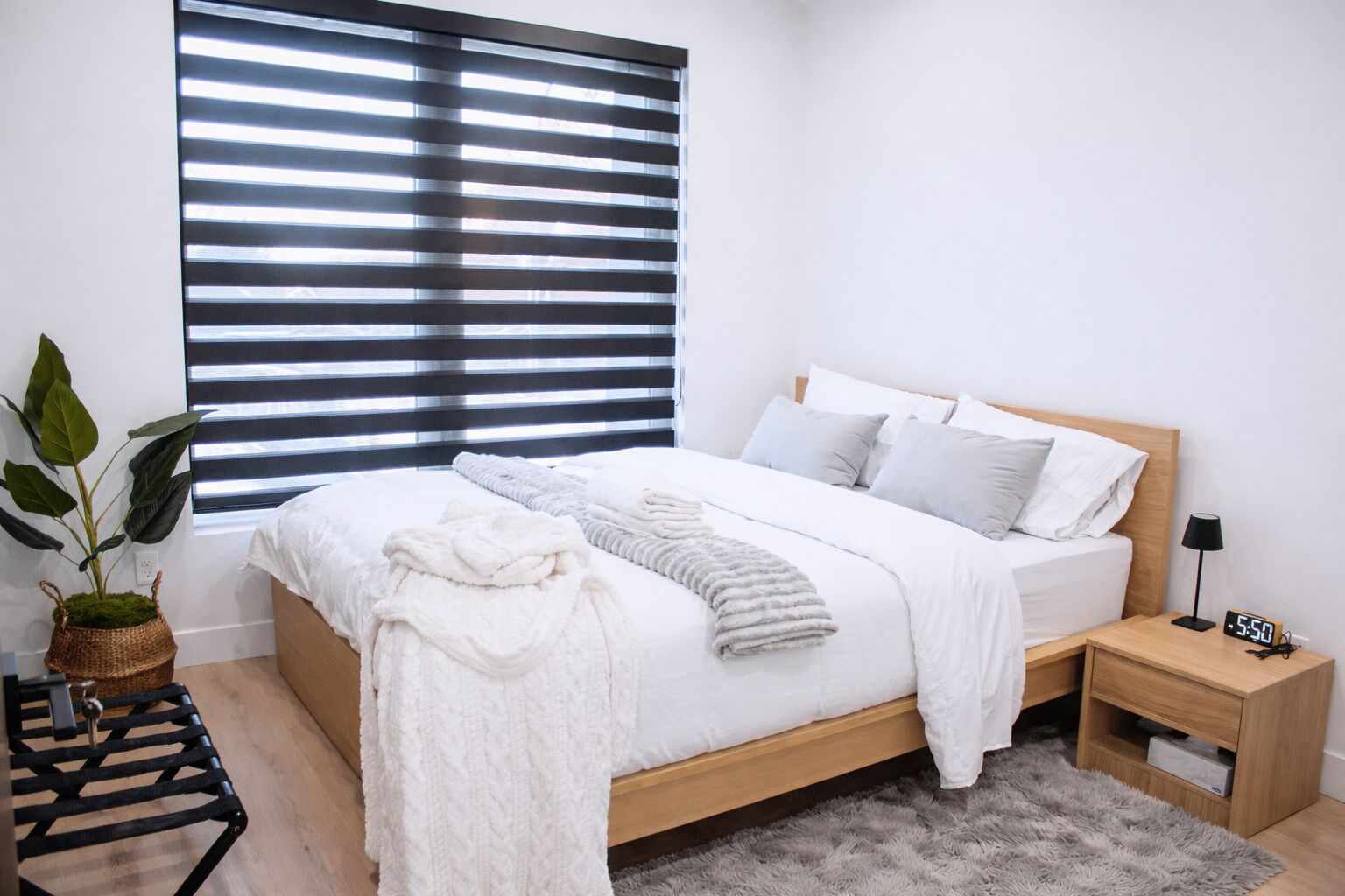 A modern bedroom with a wooden bed, white bedding, a bedside table with a lamp, and a potted plant.