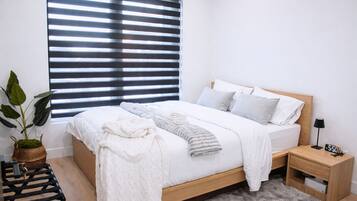 A modern bedroom with a wooden bed, white bedding, a bedside table with a lamp, and a potted plant.