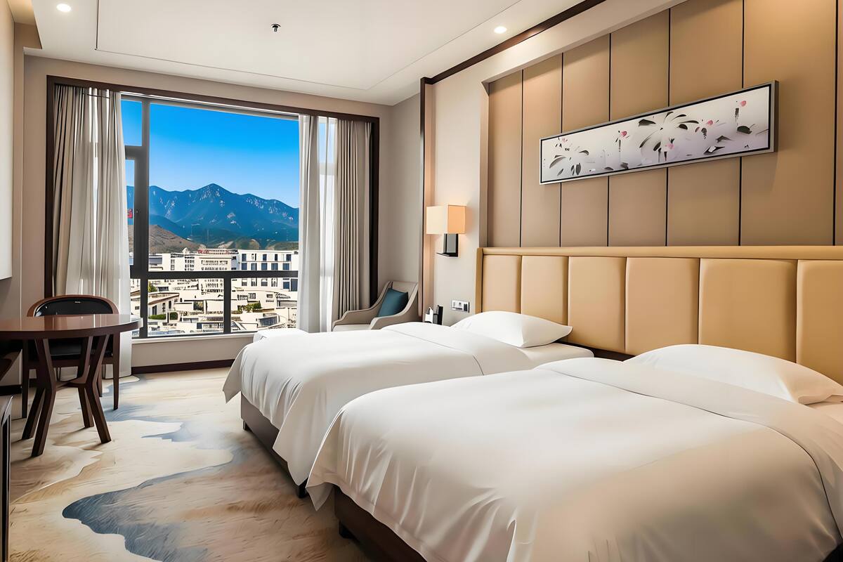 A hotel room with two beds, a desk, a chair, a large window with a mountain view, and a wall-mounted lamp.