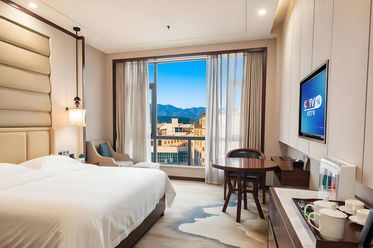 A hotel room with a bed, a desk with a chair, a TV, and a view of buildings and mountains.