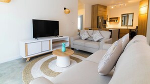 Een moderne woonkamer met een flatscreen televisie, een witte bank, een salontafel en een keuken op de achtergrond.