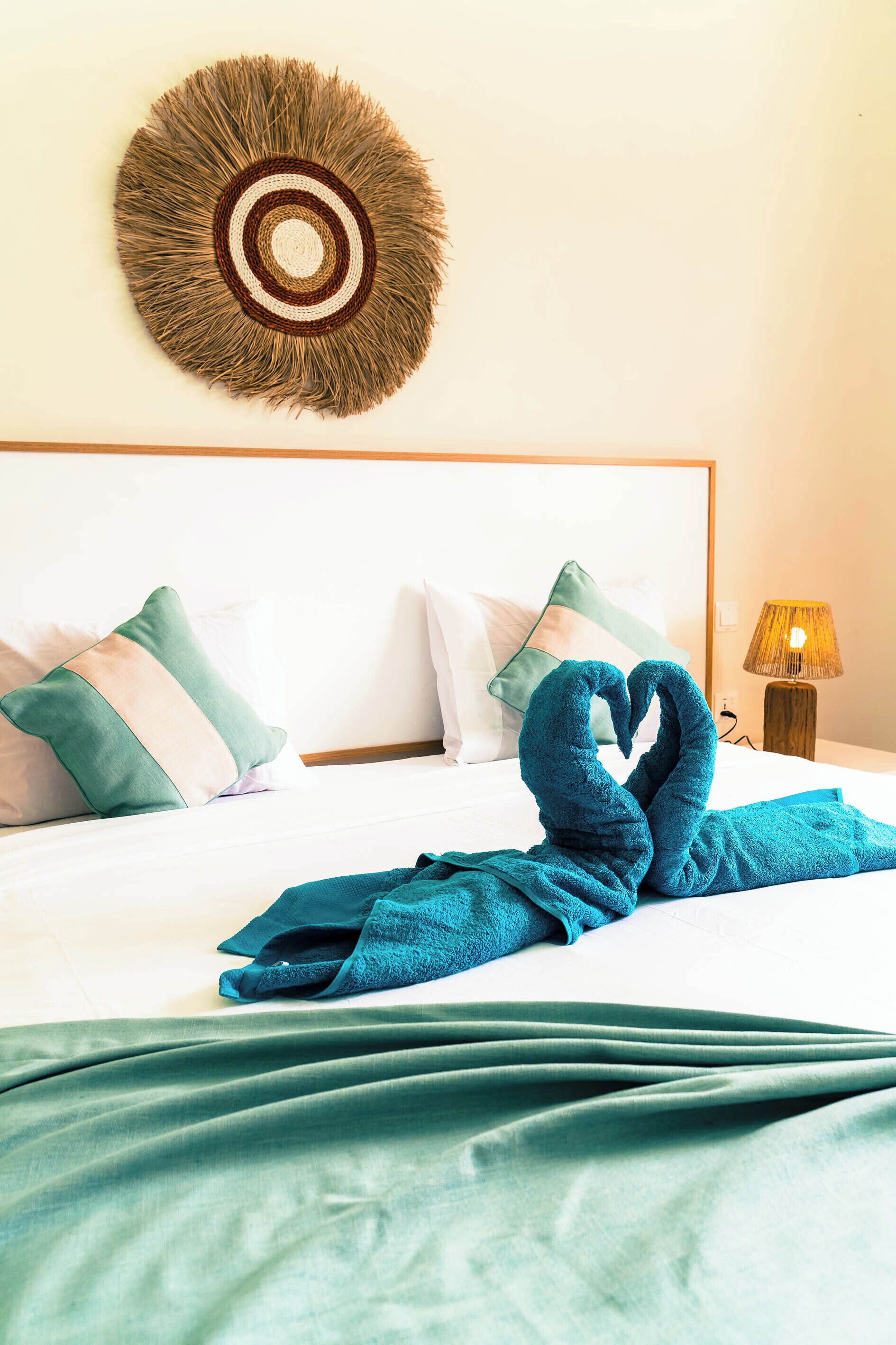 A neatly made bed with a heart-shaped towel, a decorative wall hanging, and a bedside lamp.