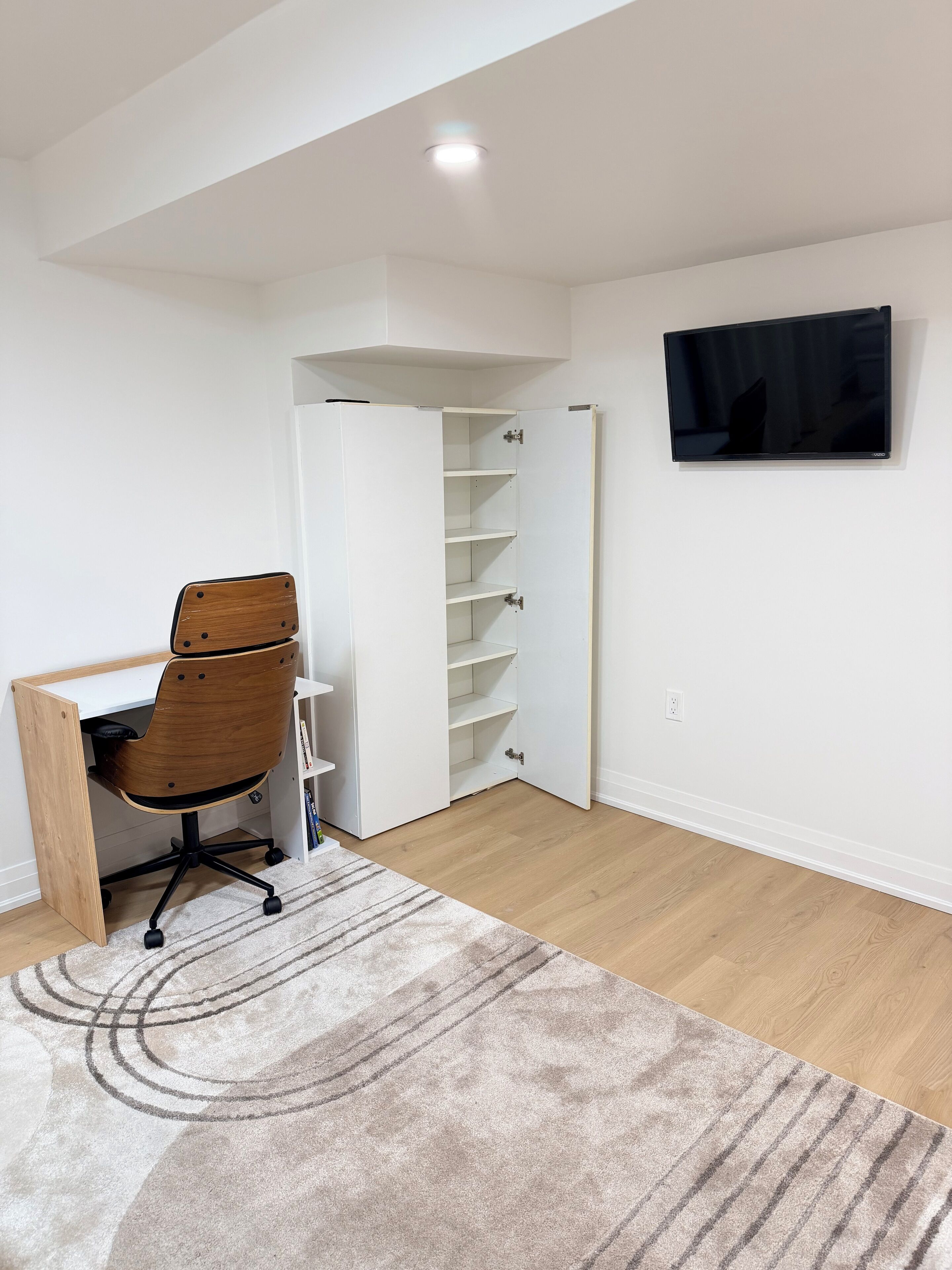 A room with a wooden desk, a chair, a white shelving unit, a flat-screen TV mounted on the wall, and a patterned rug on the floor.
