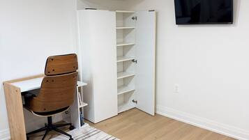 A room with a wooden desk, a chair, a white shelving unit, a flat-screen TV mounted on the wall, and a patterned rug on the floor.