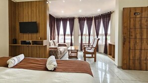 A hotel room with a bed, a TV, a seating area with a sofa and chairs, and a wooden wardrobe. - Navarathna Comforts (Madikeri)