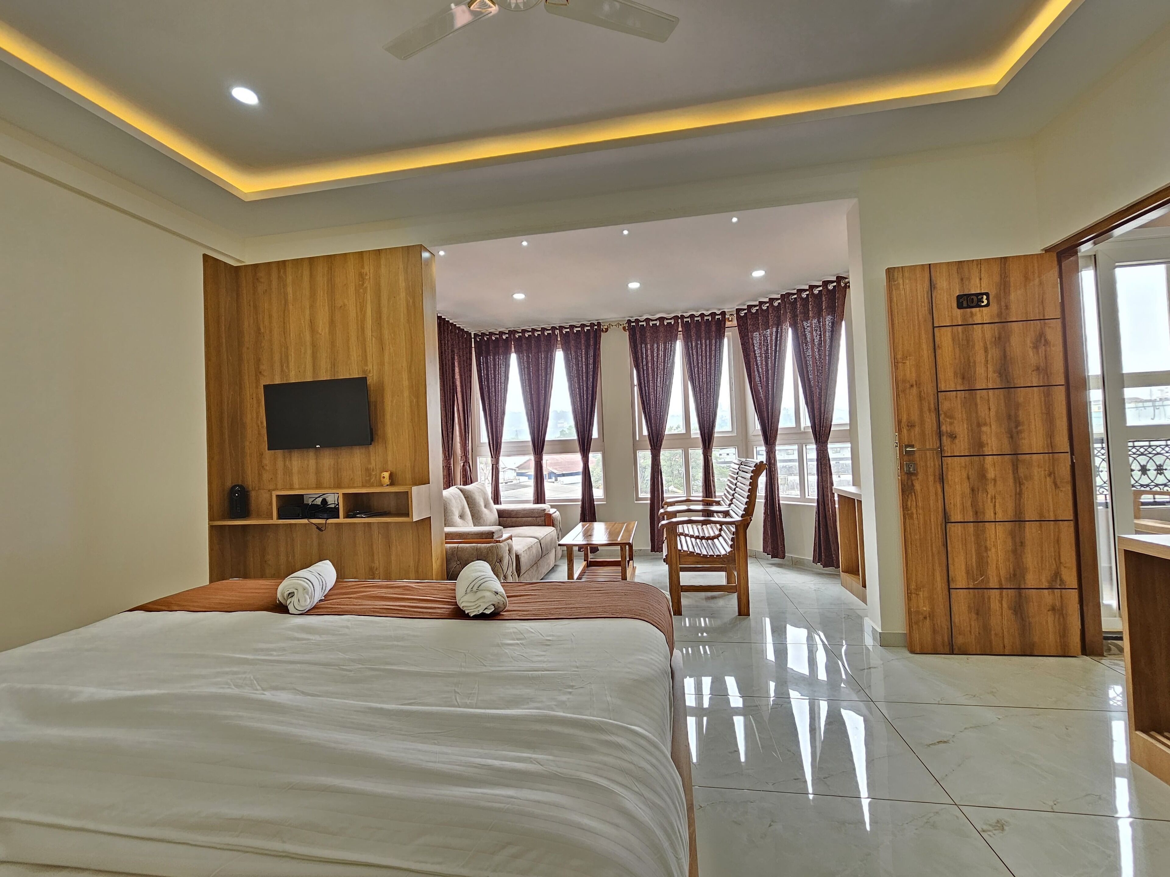 A hotel room with a bed, a TV, a seating area with a table and chairs, and a balcony with curtains.