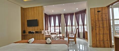 A hotel room with a bed, a TV, a seating area with a table and chairs, and a balcony with curtains.