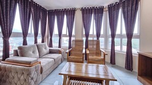A room with a sofa, two wooden chairs, a table, and large windows with curtains. - Navarathna Comforts (Madikeri)