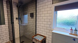 Un baño con paredes de azulejos blancos, una cabina de ducha de vidrio, un taburete pequeño de madera y una ventana con vidrio esmerilado.
