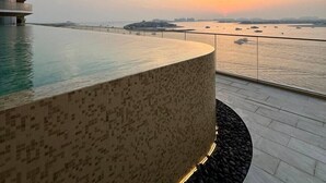 A pool with a patterned floor, a modern building with a curved facade, and a sunset view over a body of water. - Luxury 1BR with private Beach Access & Pool, Balcony with Sea View (Dubai)