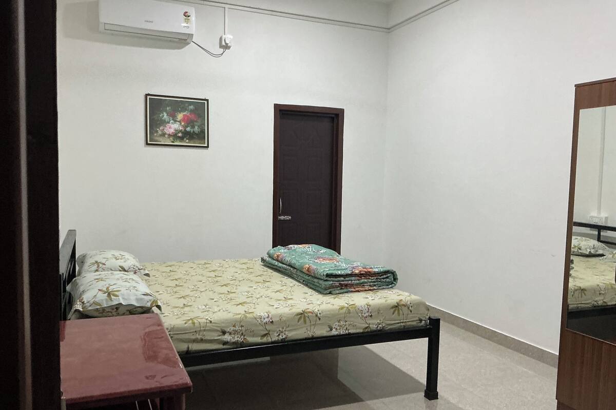 Quarto com cama, escrivaninha, cadeira, porta e ar-condicionado fixado na parede.