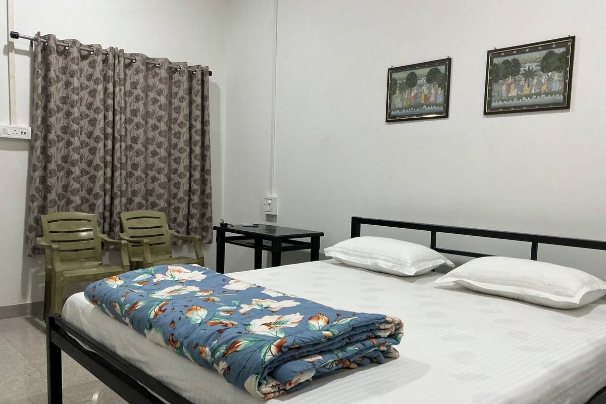 Cama bem arrumada com colcha florida, duas cadeiras, uma mesinha e um quadro na parede.
