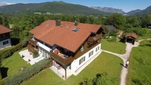 En tvåvånings chalet med ett sluttande tak, omgiven av frodig grönska och med berg i bakgrunden.