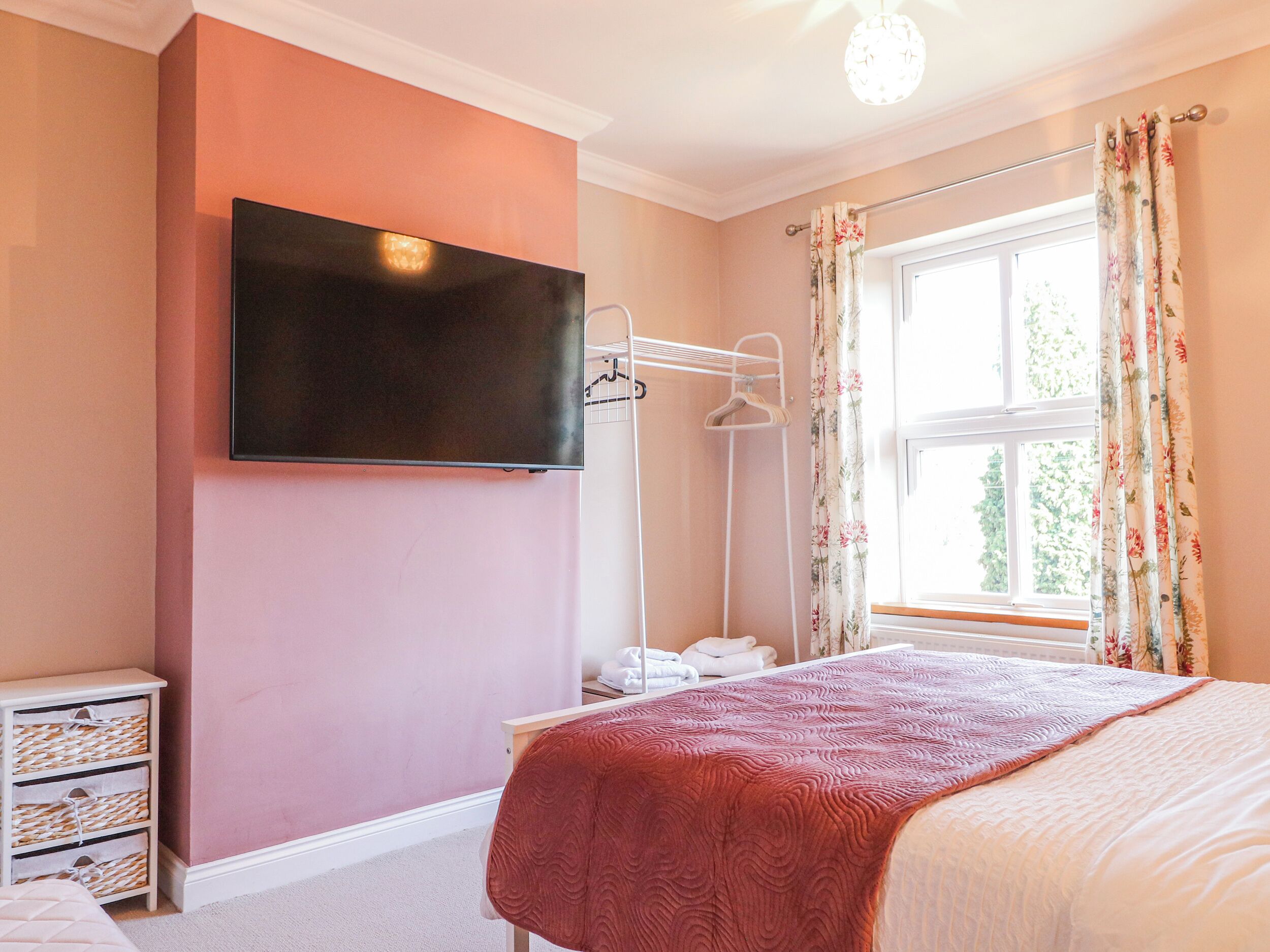A bedroom with a bed, a TV mounted on the wall, a window with curtains, and a bedside table with a lamp.