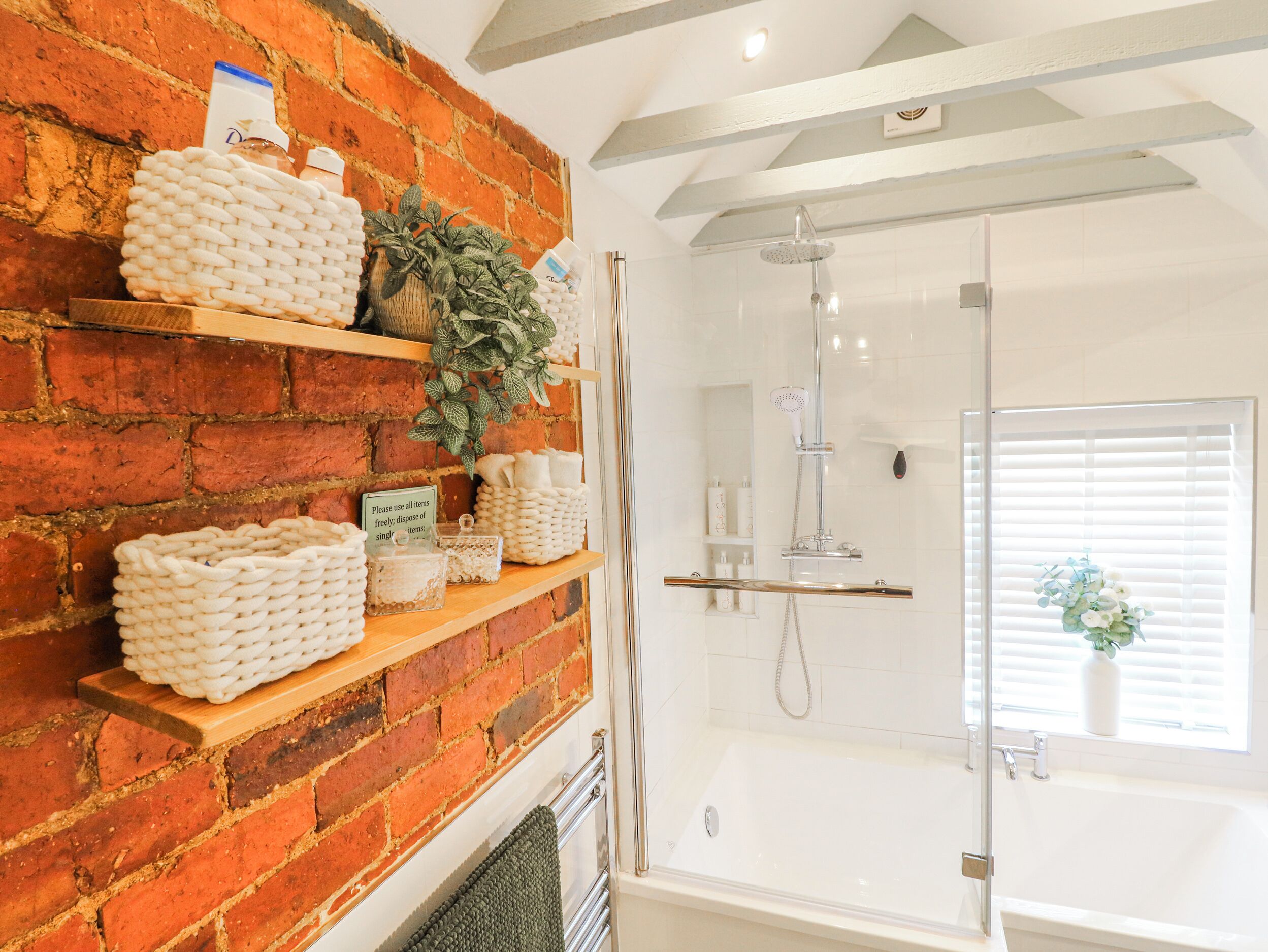 A bathroom with a brick wall, shelves with toiletries, a shower, and a window with blinds.