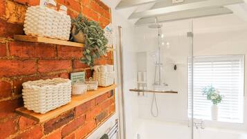 A bathroom with a brick wall, shelves with toiletries, a shower, and a window with blinds.