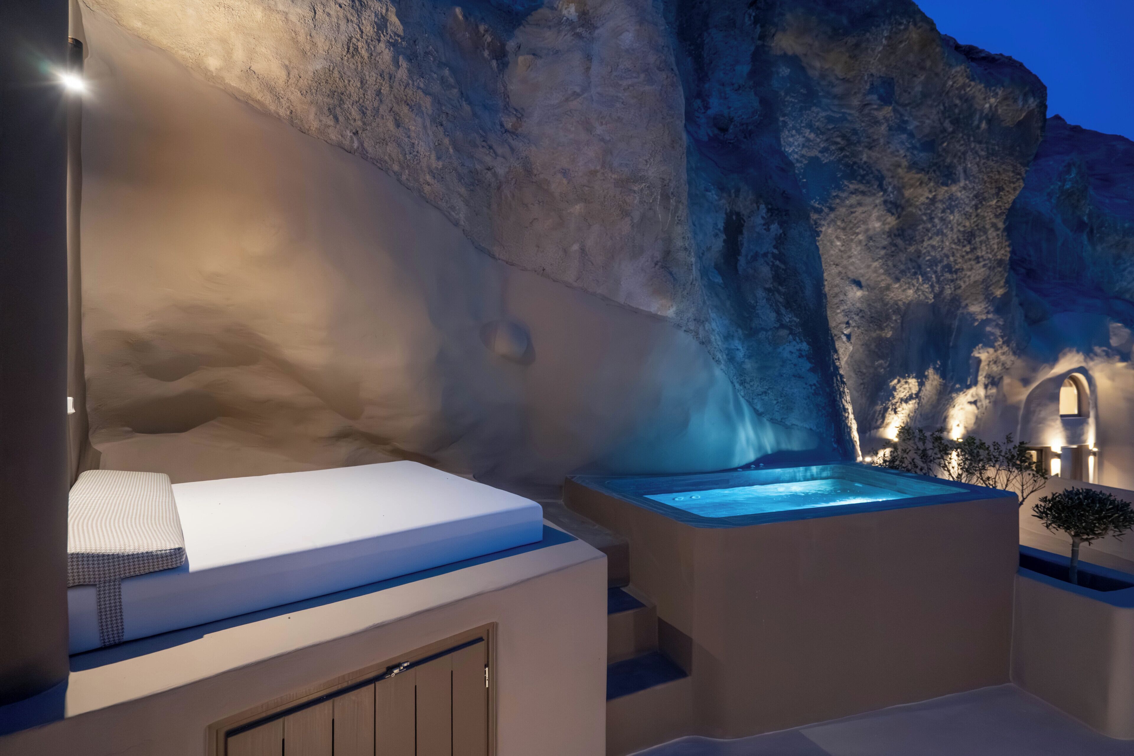 A modern outdoor relaxation area with a hot tub, a cushioned seating area, and a stone wall backdrop.