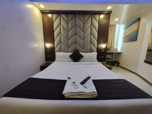 A hotel room with a bed, a nightstand, a desk, and a painting on the wall. - Hotel Rio (Mumbai)