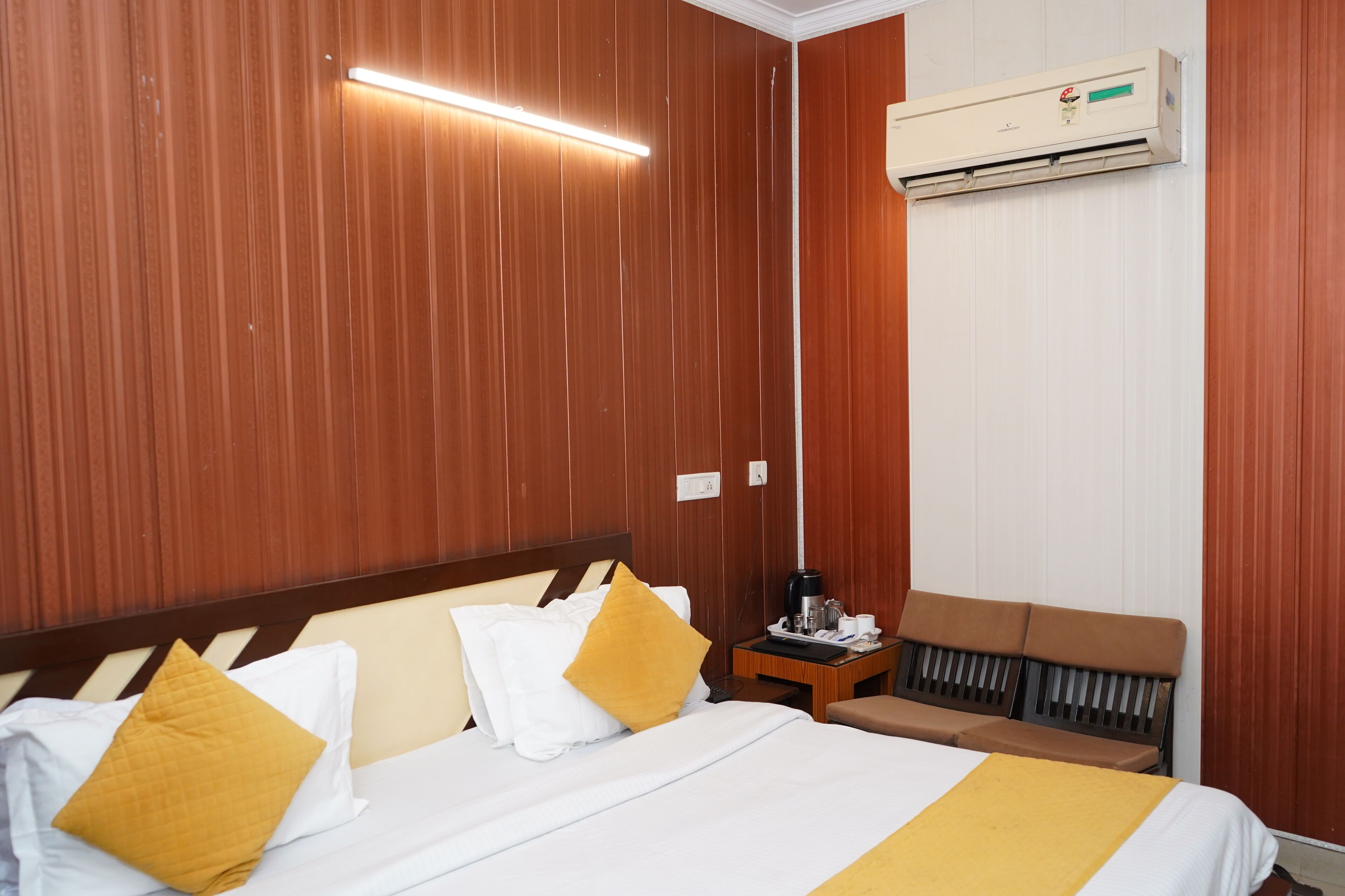A hotel room with a wooden paneled wall, a bed with white and yellow bedding, a bedside table with a coffee maker, and an air conditioning unit mounted on the wall.