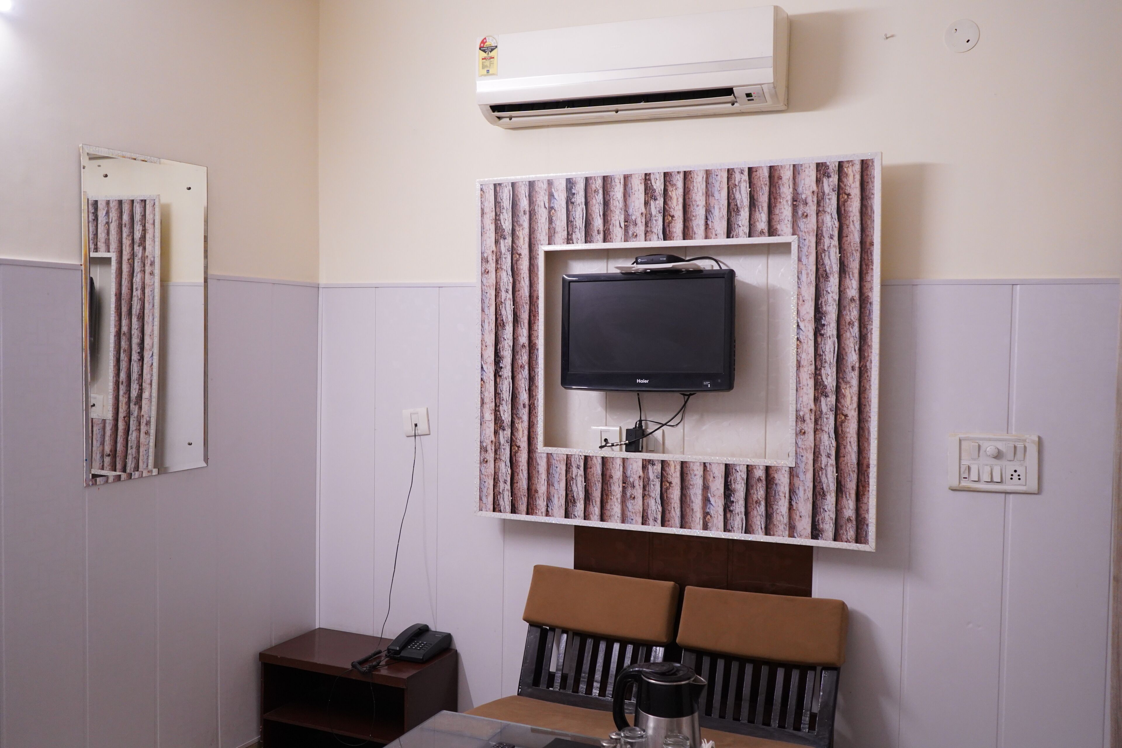 A hotel room with a mounted television, a small table with a coffee pot, a telephone, and a wall-mounted air conditioner.