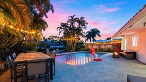 Zona de piscina con mesa y sillas de madera, rodeada de palmeras y un cielo al atardecer.