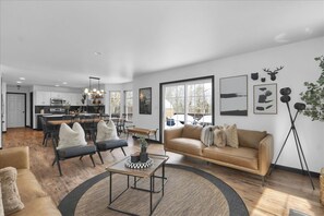 Ein modernes Wohnzimmer mit einer Couch, einem Couchtisch und Sesseln. Im Hintergrund ist eine Küche mit Schränken und einem Essbereich zu sehen.