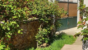 Ein hölzerner Weg führt zu einem Haus, das von viel Grün und einer Steinmauer umgeben ist, mit einem Verbotsschild für parkende Fahrzeuge.
