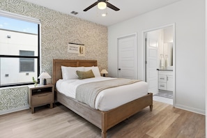 A bedroom with a wooden bed, nightstands, a window, patterned wallpaper, a ceiling fan, and a door leading to a bathroom. - Near Downtown | Modern Design + Patio + Group-Friendly | Oak Haus by AvantStay (Austin)