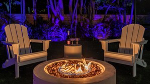 Two Adirondack chairs around a circular fire pit with a small table and two wine glasses in the center. - Enclosed Pool, Firepit, Outdoor Dining + Bar | 8 Mins to Beach | Soleil Oasis (Fort Lauderdale)