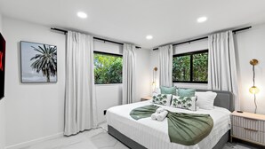 A modern bedroom with a large bed, two windows with curtains, a nightstand, a wall-mounted TV, and a framed picture. - Enclosed Pool, Firepit, Outdoor Dining + Bar | 8 Mins to Beach | Soleil Oasis (Fort Lauderdale)