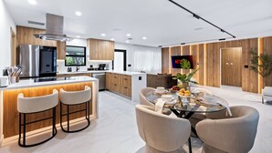A modern kitchen with a central island, bar stools, a dining table with chairs, and a TV mounted on the wall. - Enclosed Pool, Firepit, Outdoor Dining + Bar | 8 Mins to Beach | Soleil Oasis (Fort Lauderdale)