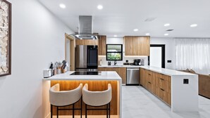 A modern kitchen with a central island, wooden cabinets, stainless steel appliances, and a large window. - Enclosed Pool, Firepit, Outdoor Dining + Bar | 8 Mins to Beach | Soleil Oasis (Fort Lauderdale)